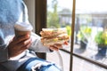 A woman holding and eating whole wheat sandwich and coffee in the morning