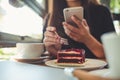 Closeup image of a woman holding , using and looking at smart phone while eating a cake with white coffee cups and laptop on woode Royalty Free Stock Photo