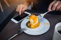 Closeup image of a woman using smartphone while cutting and eating an orange cake with fork on white plate on wooden table Royalty Free Stock Photo