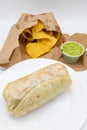 Large Burrito in front of a Bag of Tortilla Chips and a Cup of Guacamole on a White Background Royalty Free Stock Photo