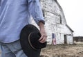 Closeup of man holding black cowboy hat standing in front of aandoned old barn on farm with American flag Royalty Free Stock Photo