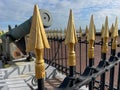 Closeup of metal fence with wooden heads and an old cannon behind with blue sky