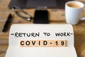 Closeup on notebook over wood table background, focus on wooden blocks with letters making Covid-19 Return To Work writing