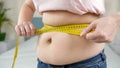 Closeup of obese woman measuring her big belly with yellow measuring tape. Concept of dieting, unhealthy lifestyle