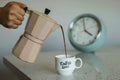 Closeup of a person pouring coffee from a moka pot with an alarm clock in the background