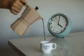 Closeup of a person pouring coffee from a moka pot with an alarm clock in the background Royalty Free Stock Photo