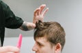 Closeup photo of a male hairdresser trimming a young man against a gray wall background holding a comb in his hand. Barber creates