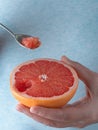 Closeup, photo of a woman holding half of fresh ripe red grapefruit in her hand. Spoon with a slice of grapefruit in hand. Blue