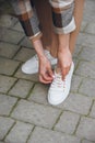 Closeup photo of young woman wearing checkered long coat, and beige pants . Lady posing on city street. Women ties laces