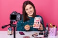 Closeup picture of young positive smiling European beauty vlogger sitting at table against camera and presents eyeshadow palette,