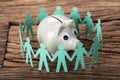 Piggybank Surrounded By Paper People Holding Hands On Wood