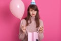 Closeup portrait of beautiful disappointed girl with gift package and balloon, wearing pink sweater and birthday hat, posing Royalty Free Stock Photo
