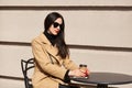 Closeup portrait of beautiful stylish model sitting at brown wooden table, drinking coffee, looks calm. Fashionable young lady