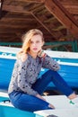 Closeup portrait of beautiful young blond girl in jeans and sneakers sitting on blue boat on beach. Royalty Free Stock Photo