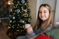 Closeup portrait of cheerful adorable little girl holding in hands mug with hot cocoa sitting on sofa by Christmas tree Royalty Free Stock Photo