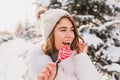 Closeup portrait cute funny girl in white woolen hat having fun with pink heart lollypop on street. Pretty young woman Royalty Free Stock Photo