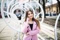 Closeup portrait of enjoyed brunette girl speaking on phone on street near the modern bench. She wears pink jacket, smiling to sid Royalty Free Stock Photo