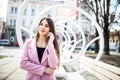 Closeup portrait of enjoyed brunette girl speaking on phone on street near the modern bench. She wears pink jacket, smiling to sid Royalty Free Stock Photo