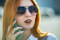 Closeup portrait of fashionable young redhead woman in sunglasses talking on cell phone outdoors in summer Royalty Free Stock Photo