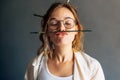 Closeup portrait of a funny beautiful female artist in eyeglasses holding with her lips a painting brush in her workshop. Pretty Royalty Free Stock Photo