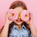 Closeup portrait happy cute little girl in blue shirt holding donuts on eyes, having fun to camera isolated on pink Royalty Free Stock Photo