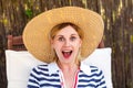 Closeup portrait of happy laughing unexpectedly young adult woman in hat and dress sitting on cozy deckchair and looking at camera