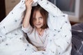 Closeup portrait of happy little girl sitting on bed under blanket and posing in room of her flat holds plaid under her head, Royalty Free Stock Photo