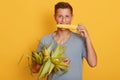 Closeup portrait of happy young student guy biting maize and holding other corns in hands, stands against yellow wall, attractive