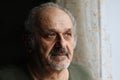 Unhappy old man indoors: senior man with grey beard and moustache standing near window. Loneliness, aged people problems concept