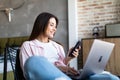 Portrait of smiling young beautiful woman talking on smartphone, working on laptop and sitting on chair at home Royalty Free Stock Photo