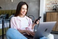 Portrait of smiling young beautiful woman talking on smartphone, working on laptop and sitting on chair at home Royalty Free Stock Photo