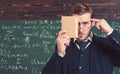 Closeup portrait of student holding book in front of his face and pressing finger over forehead. Thoughtful intelligent