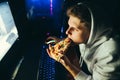 Closeup portrait of a young gamer playing video games at home on a computer, eating a slice of pizza, looking intently at the