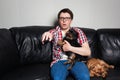 Closeup portrait, young man in the red shirt, sitting on black leather couch with two dogs, watching TV, holding remote, surprised Royalty Free Stock Photo