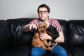 Closeup portrait, young man in the red shirt, sitting on black leather couch with two dogs, watching TV, holding remote, surprised Royalty Free Stock Photo