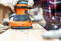 Closeup professional carpenter hand grinding raw wood plank with orbital sander electric machine in carpentry diy workshop. Detail