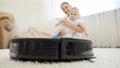 Closeup of robot vacuum cleaner riding past smiling mother with baby son at livign room
