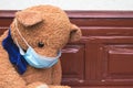 Closeup of sad teddy bear toy wearing disposable medical protective face mask sitting on vintage brown bench. Stay at home and Royalty Free Stock Photo