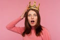 Closeup shocked bossy woman with brown hair in pink sweater adjusting golden crown on head looking at camera with big eyes and