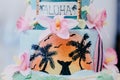 Closeup shot of a cake with Hawaiian decorations with pink flowers, palms and the word ALOHA