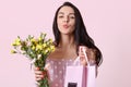 Closeup shot of good looking dark haired young woman keeps lips folded, holds gift bag and flowers, gives present to friend, poses