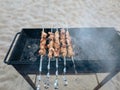 Closeup of some grilled meat skewers in a barbecue standing on the sand by the sea