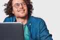 Closeup studio shot of a young smiling man with curly hair sitting at table working at laptop and dreaming. Royalty Free Stock Photo