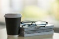 Closeup of takeaway papercup of hot coffee with stack of books and reading glasses on wooden table