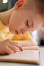 Closeup vertical shot of cute pupil schoolboy reading paper book following lines with finger along page sitting at desk Royalty Free Stock Photo