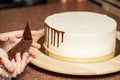 Closeup of a woman decorating white smooth cake's edges with chocolate in a pipe bag, in the kitchen