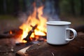 Closeup of white enamel cup outdoors in camping, with burning campfire on background.