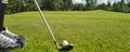 Closeup of white golf ball and club. Human feet on green grass