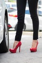 Closeup womans legs wearing black jeans and red stilettos standing outside car door, other cars background, female