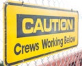 Closeup of a yellow [Caution Crews Working Below] sign on a chain-link fence under the sunlight